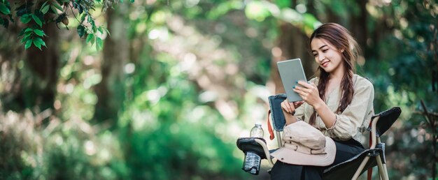 Junge hübsche Frau, die auf einem Stuhl sitzt und Tablet-Videoanrufe nutzt, während sie im Naturpark-Kopienraum campt