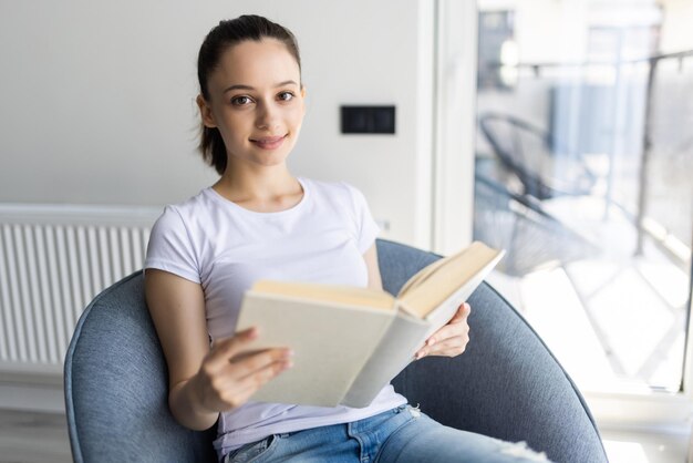 Junge hübsche Frau, die ein Buch liest und zu Hause auf einem bequemen Stuhl sitzt