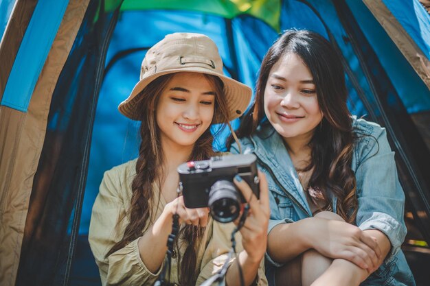 Junge hübsche Frau, die ein Foto mit der Kamera macht und ihrer Freundin ein Bild zeigt, während sie im Campingzelt im Wald sitzt. Junge asiatische Gruppenfrauen reisen im Freien