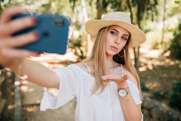 Junge hübsche Frau, die ein Selfie in einem stilvollen Sommerhut in einem Park nimmt.