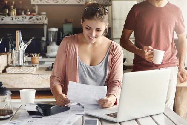 Junge hübsche Frau, die eine Brille auf ihrem Kopf trägt, lächelt glücklich, während sie Dokument liest, das sagt, dass Bank ihren Hypothekenantrag genehmigte