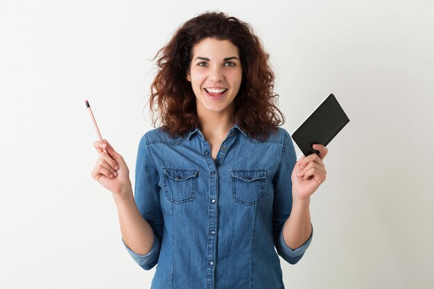 Junge hübsche Frau, die Hände mit Notizbuch und Bleistift hochhält, lächelnd, überraschter Gesichtsausdruck, lockiges Haar, positive Emotion, glücklich, isoliert, blaues Jeanshemd, Student, Bildung