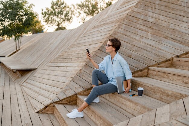 Junge hübsche Frau, die mit Laptop in der modernen Parkstraße mit Smartphone sitzt