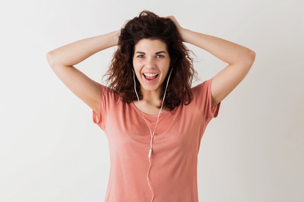 Junge hübsche Frau, die Musik auf Kopfhörern hört, Hände an ihrem lockigen Haar hochhält, lacht, lustiger Gesichtsausdruck, positive Emotion, glücklich, isoliert, rosa T-Shirt, Student