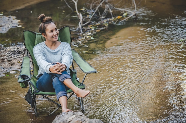 Junge hübsche Frau, die zur Entspannung auf einem Campingstuhl im Bach sitzt, lächelt sie im Naturwald, während sie einen Campingausflug mit Glückskopierraum macht