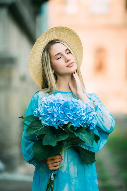 Junge hübsche Frau im blauen Kleid, die die Blumen der Hydragea in der Sommerstraße hält