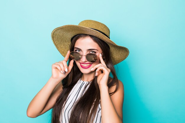 Junge hübsche Frau im Sommerkleidungshut und in der Sonnenbrille auf türkisfarbenem Hintergrund