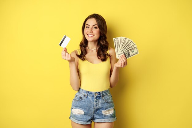 Junge hübsche Frau im Sommeroutfit mit Kreditkarte und Geld, Bargeld in der Hand, vor gelbem Hintergrund stehend.