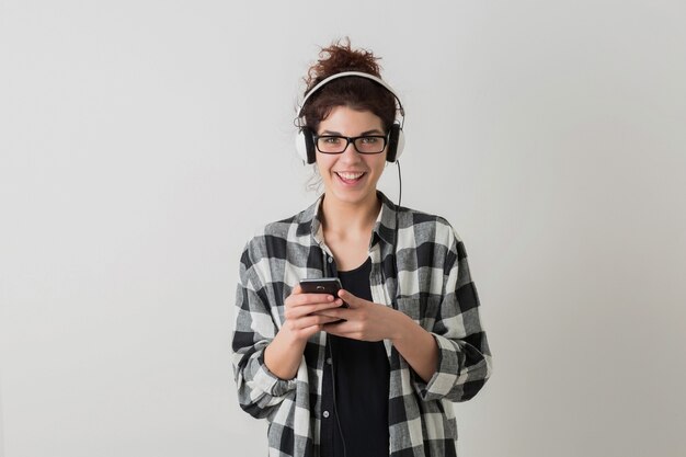 Junge hübsche Frau in der Brille, mit Smartphone, emotional, lachend, positiv, glücklich, Musik auf Kopfhörern hörend, isoliert, kariertes Hemd, Hipster-Stil, Student, in der Kamera schauend