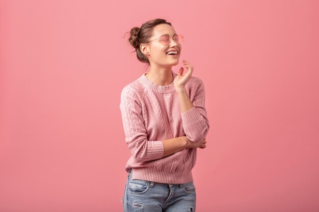 Junge hübsche Frau mit aufrichtigem offenem Lächeln im rosa Pullover und in der Sonnenbrille lokalisiert auf rosa Studiohintergrund