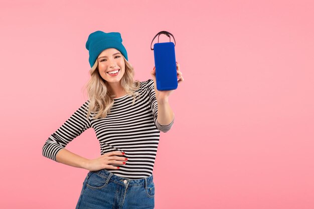 Junge hübsche Frau mit drahtlosem Lautsprecher, die Musik mit gestreiftem Hemd und blauem Hut hört, lächelt glücklich positive Stimmung und posiert isoliert auf rosafarbenem Hintergrund
