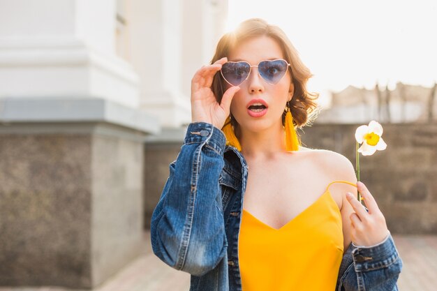 Junge hübsche Frau mit überraschtem Gesichtsausdruck, emotionaler, schockierter Emotion, tragen stilvolle Kleidung, Jeansjacke, gelbes Oberteil, Halteblume, sonniger Sommer, trendige lustige blaue Sonnenbrille