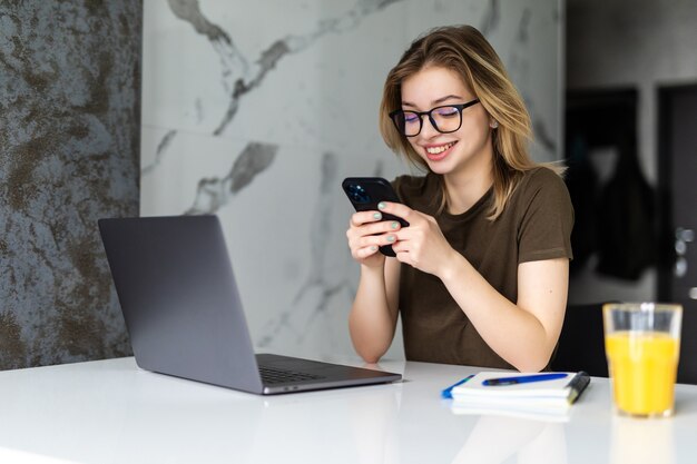 Junge hübsche Frau sitzt mit einem Laptop am Küchentisch und benutzt ein Handy