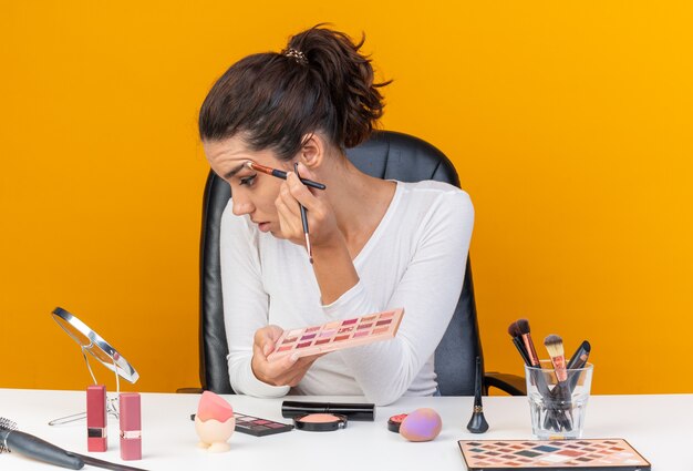 Junge hübsche kaukasische Frau, die am Tisch mit Make-up-Tools sitzt, die Lidschatten-Palette hält und Lidschatten mit Make-up-Pinsel auf den Spiegel aufträgt