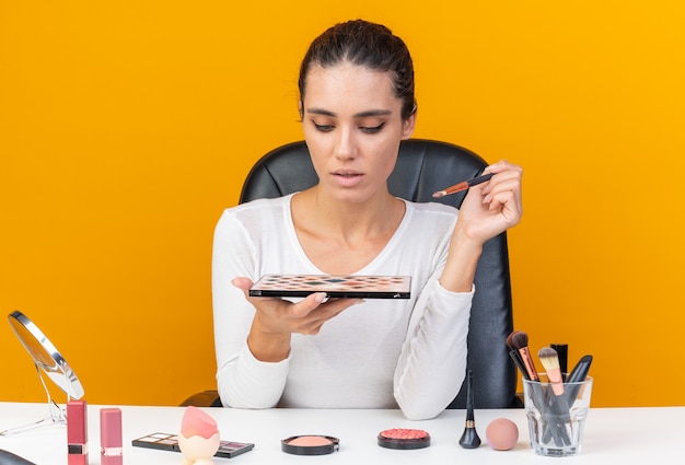 Junge hübsche kaukasische Frau, die am Tisch mit Make-up-Tools sitzt und Make-up-Pinsel und Lidschatten-Palette hält