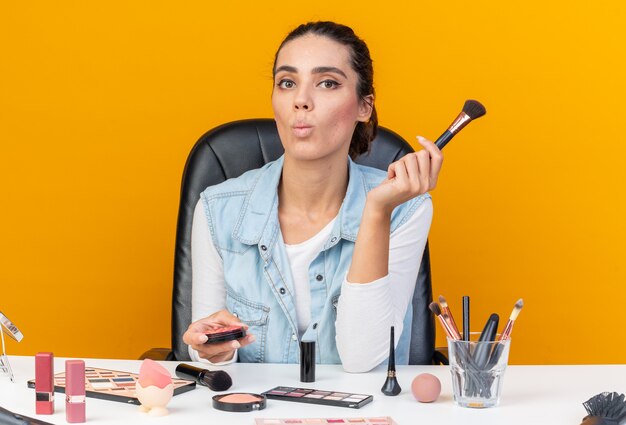 Junge hübsche kaukasische Frau, die am Tisch mit Make-up-Werkzeugen sitzt, die Fischgesicht mit Rouge und Make-up-Pinsel einzeln auf oranger Wand mit Kopierraum machen