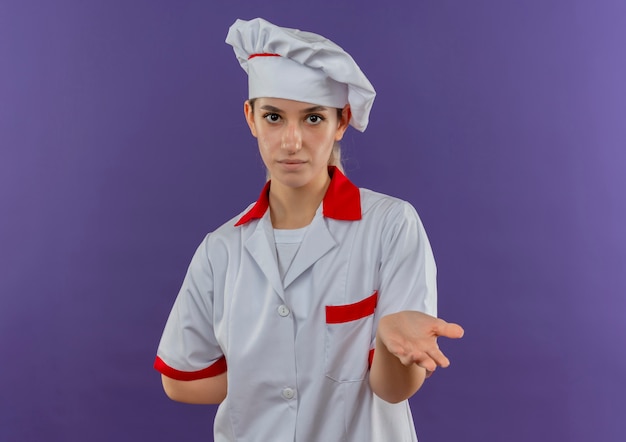 Junge hübsche Köchin in Kochuniform zeigt leere Hand, die eine andere Hand hinter ihren Rücken legt und schaut