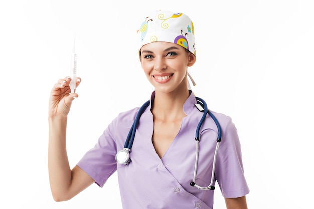 Junge hübsche lächelnde Ärztin in Uniform mit Phonendoskop am Hals, die Spritze in der Hand hält, während sie glücklich vor weißem Hintergrund in die Kamera blickt