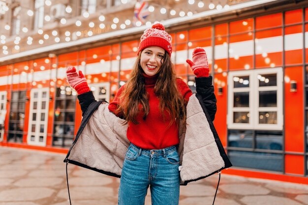 Junge hübsche lächelnde aufgeregte glückliche Frau in den roten Handschuhen und in der gestrickten Mütze, die Wintermantel trägt, der in der Stadtweihnachtsstraße, Modetrend der warmen Kleidungsstil geht