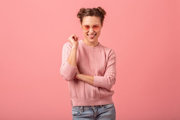 Junge hübsche lächelnde Frau mit lustigem Gesichtsausdruck, der Zunge in rosa Pullover und Sonnenbrille lokalisiert auf rosa Studiohintergrund zeigt