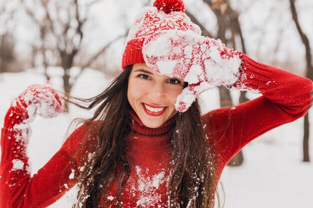 Junge hübsche lächelnde glückliche Frau in den roten Handschuhen und im Hut, die den gestrickten Pullover tragen, der im Park im Schnee, warme Kleidung, Spaß hat und lange Haare winkt