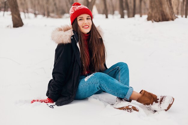 Junge hübsche lächelnde glückliche Frau in den roten Handschuhen und in der gestrickten Mütze, die Wintermantel trägt, der auf Schnee im Park, warme Kleidung sitzt