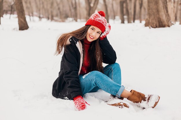 Junge hübsche lächelnde glückliche Frau in den roten Handschuhen und in der gestrickten Mütze, die Wintermantel trägt, der auf Schnee im Park, warme Kleidung sitzt