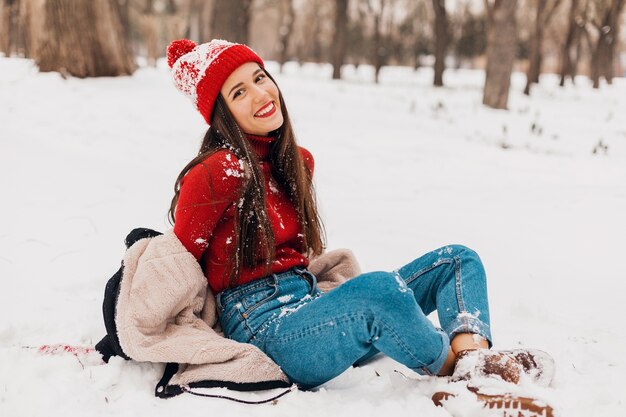 Junge hübsche lächelnde glückliche Frau in den roten Handschuhen und in der gestrickten Mütze, die Wintermantel trägt, der auf Schnee im Park, warme Kleidung sitzt