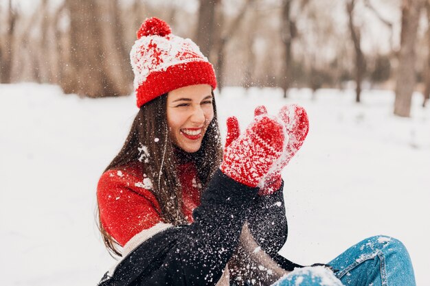 Junge hübsche lächelnde glückliche Frau in den roten Handschuhen und in der gestrickten Mütze, die Wintermantel trägt, der auf Schnee im Park, warme Kleidung sitzt