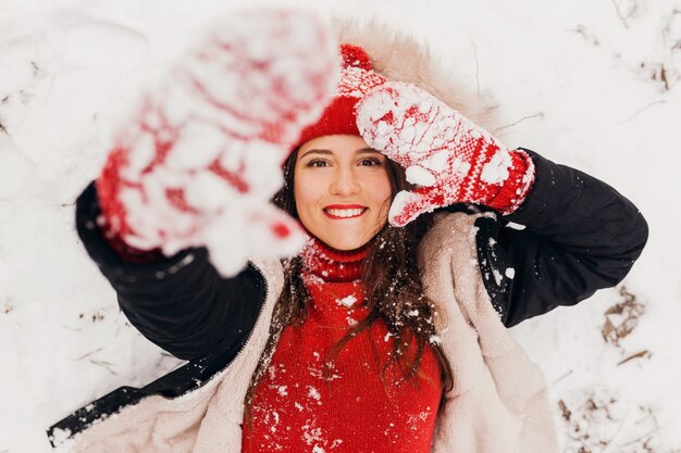 Junge hübsche lächelnde glückliche Frau in den roten Handschuhen und in der gestrickten Mütze, die Wintermantel trägt, der im Park im Schnee, warme Kleidung, Ansicht von oben liegt