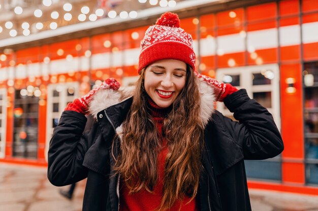 Junge hübsche lächelnde glückliche Frau in den roten Handschuhen und in der gestrickten Mütze, die Wintermantel trägt, der in der Stadtweihnachtsstraße geht, warmer Kleidungsstil-Modetrend