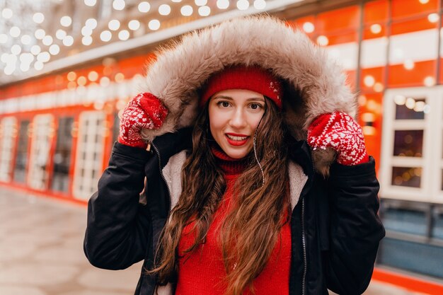 Junge hübsche lächelnde glückliche Frau in den roten Handschuhen und in der gestrickten Mütze, die Wintermantel trägt, der in der Stadtweihnachtsstraße geht, warmer Kleidungsstil-Modetrend