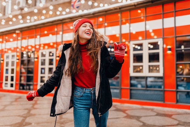 Junge hübsche lächelnde glückliche Frau in den roten Handschuhen und in der gestrickten Mütze, die Wintermantel trägt, der in Stadtstraße, warme Kleidung geht
