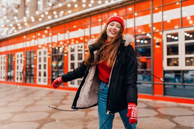Junge hübsche lächelnde glückliche Frau in den roten Handschuhen und in der gestrickten Mütze, die Wintermantel trägt, der in Stadtstraße, warme Kleidung geht