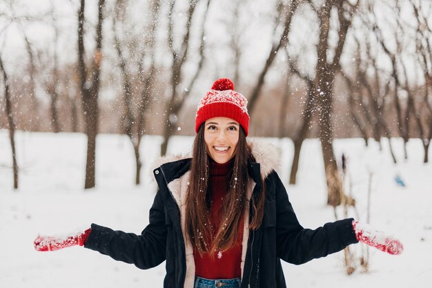 Junge hübsche lächelnde glückliche Frau in den roten Handschuhen und in der gestrickten Mütze, die Wintermantel tragen, im Park am schneebedeckten Tag, warme Kleidung