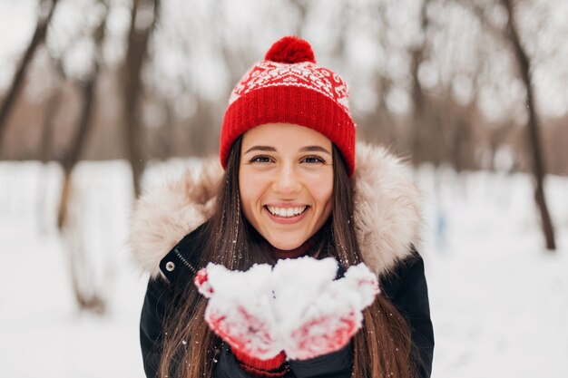 Junge hübsche lächelnde glückliche Frau in den roten Handschuhen und in der gestrickten Mütze, die Wintermantel tragen, im Park gehen, Schnee weht