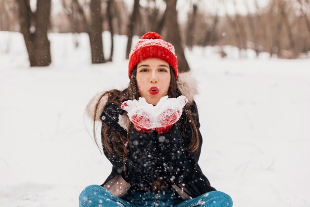 Junge hübsche lächelnde glückliche Frau in den roten Handschuhen und in der gestrickten Mütze, die Wintermantel tragen, im Park spazieren gehen, mit Schnee in der warmen Kleidung spielen