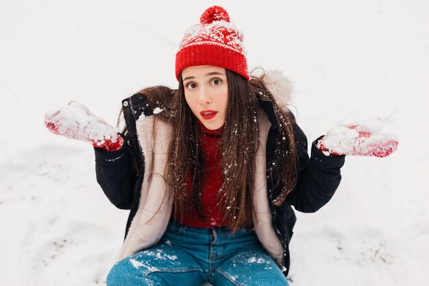 Junge hübsche lächelnde glückliche Frau in den roten Handschuhen und in der gestrickten Mütze, die Wintermantel tragen, im Park spazieren gehen, mit Schnee in der warmen Kleidung spielen