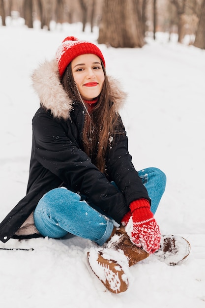 Junge hübsche lächelnde glückliche Frau in den roten Handschuhen und in der gestrickten Mütze, die Wintermantel tragen, im Park spazieren gehen, mit Schnee in der warmen Kleidung spielen