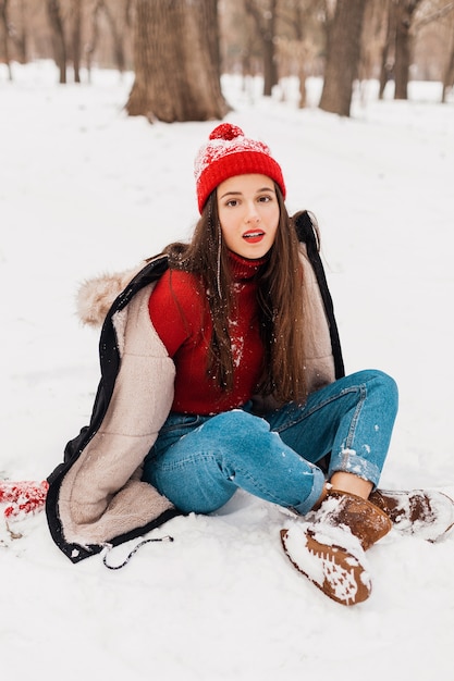 Junge hübsche lächelnde glückliche Frau in den roten Handschuhen und in der gestrickten Mütze, die Wintermantel tragen, im Park spazieren gehen, mit Schnee in der warmen Kleidung spielen