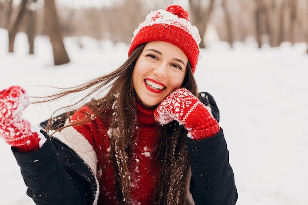 Junge hübsche lächelnde glückliche Frau in den roten Handschuhen und in der gestrickten Mütze, die Wintermantel tragen, im Park spazieren gehen, mit Schnee in der warmen Kleidung spielen