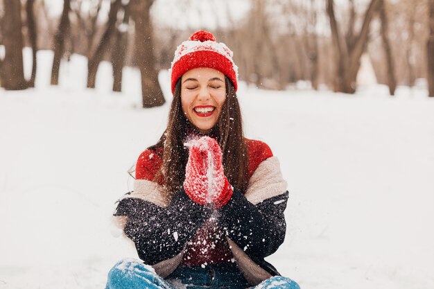 Junge hübsche lächelnde glückliche Frau in den roten Handschuhen und in der gestrickten Mütze, die Wintermantel tragen, im Park spazieren gehen, mit Schnee in der warmen Kleidung spielen