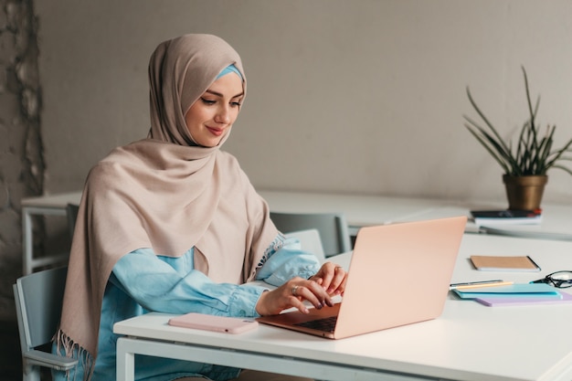 Junge hübsche moderne muslimische Frau im Hijab, die am Laptop im Büroraum arbeitet, Bildung online