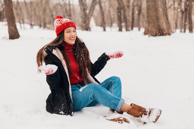 Junge hübsche offen lächelnde glückliche Frau in den roten Handschuhen und in der gestrickten Mütze, die schwarzen Mantel trägt, der im Park im Schnee spielt, warme Kleidung, Spaß hat