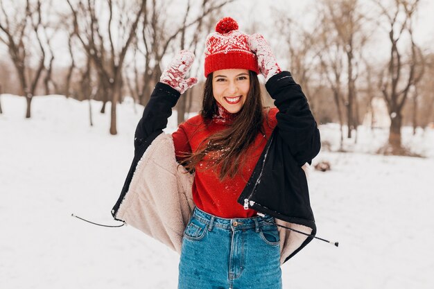 Junge hübsche offen lächelnde glückliche Frau in den roten Handschuhen und in der gestrickten Mütze, die schwarzen Mantel trägt, der im Park im Schnee spielt, warme Kleidung, Spaß hat