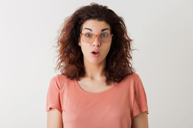 Junge hübsche stilvolle Frau in der Brille, lockiges Haar, schockiert, überraschter Gesichtsausdruck, lustige Emotion, isoliert, rosa T-Shirt