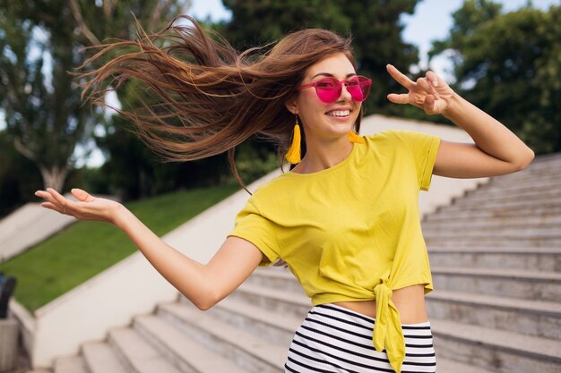 Junge hübsche stilvolle lächelnde Frau, die Spaß im Stadtpark hat, positiv, emotional, tragendes gelbes Oberteil, gestreiften Minirock, rosa Sonnenbrille, Sommerart-Modetrend, langes Haar, Friedenszeichen zeigend