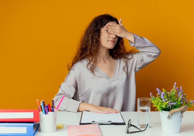 Junge hübsche weibliche Büroangestellte sitzen am Schreibtisch mit Bürowerkzeugen bedeckten Augen mit der Hand lokalisiert auf Orange