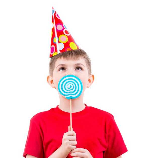Junge im roten T-Shirt und im Partyhut, die farbige Süßigkeiten essen - lokalisiert auf Weiß.