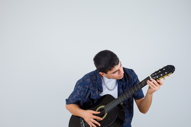 Junge im T-Shirt, das guiter spielt, während Aganist sitzt und selbstbewusst, Vorderansicht schaut.